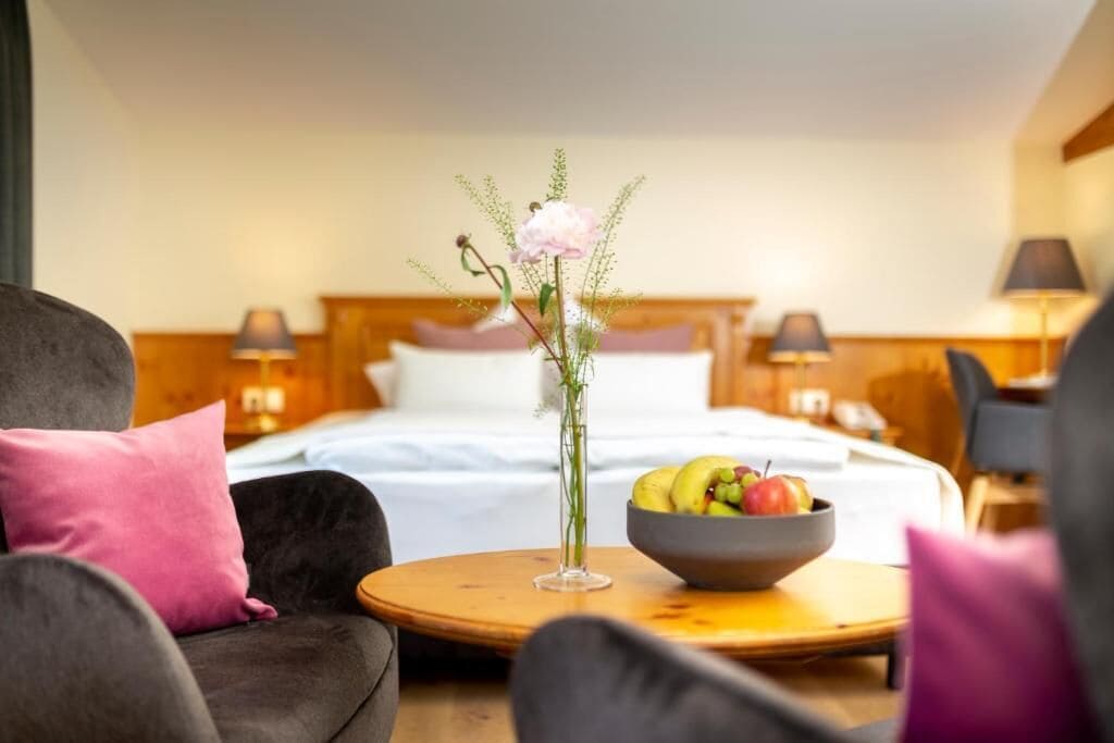 Ein Hotelzimmer mit einem Bett, einem runden Holztisch mit einer Obstschale, einer Blumenvase und einem Stuhl mit einem rosafarbenen Kissen.