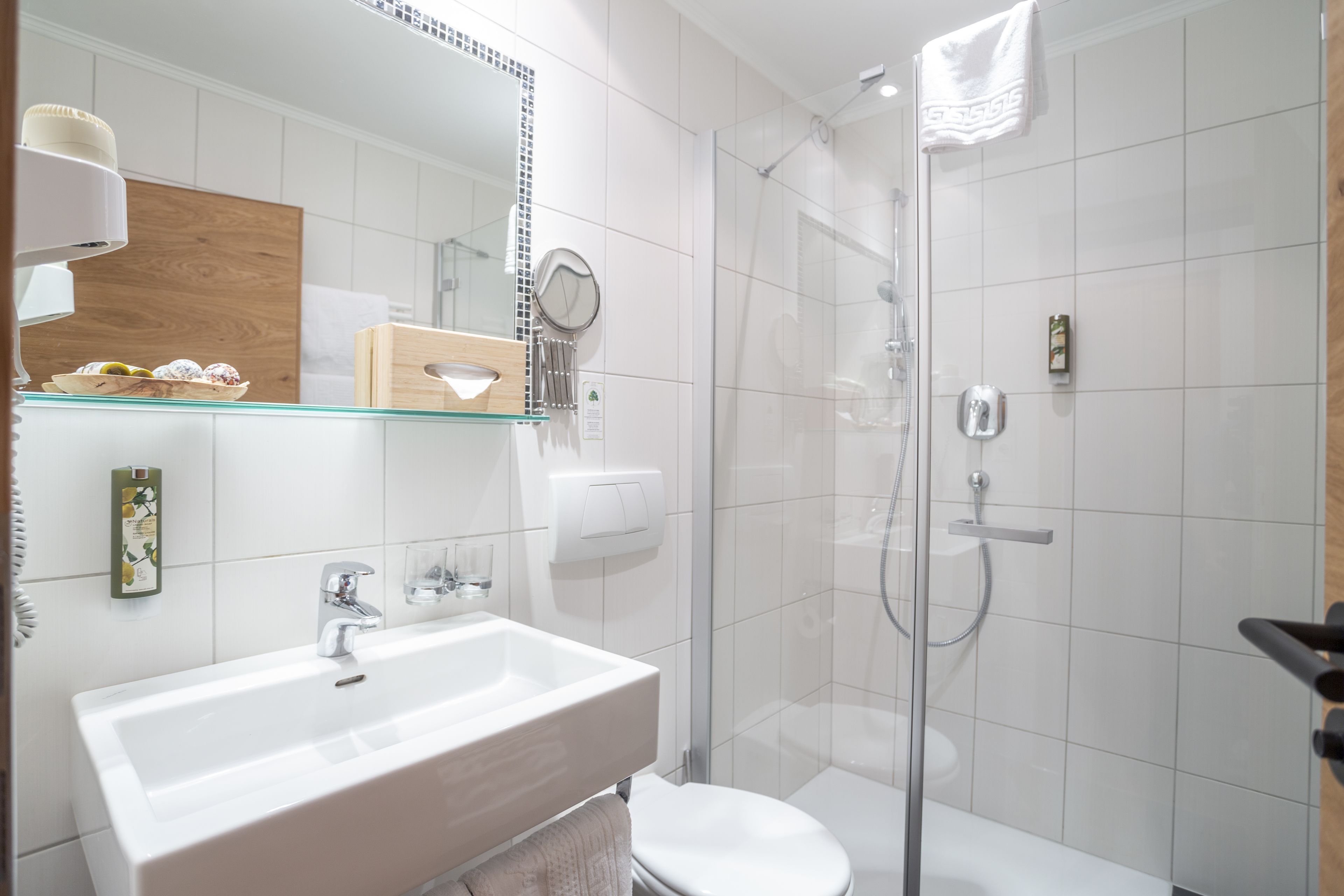 Un baño moderno con ducha de vidrio, un lavabo blanco y un estante de madera con artículos de aseo.