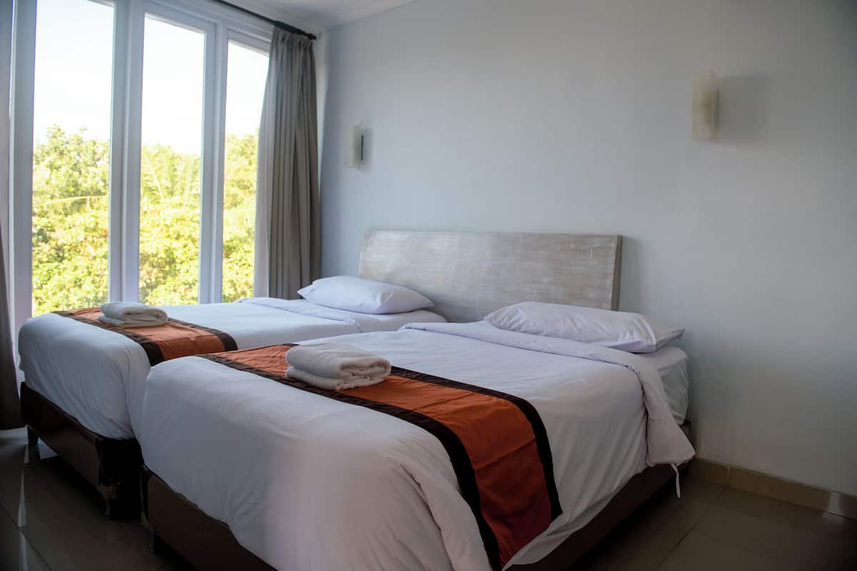 A hotel room with two beds, each with white linens and brown accents, large windows with curtains, and a view of greenery outside.