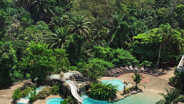 Ein Resort mit großem Pool, einem Wasserfall und einer Liegewiese, umgeben von üppiger Vegetation.