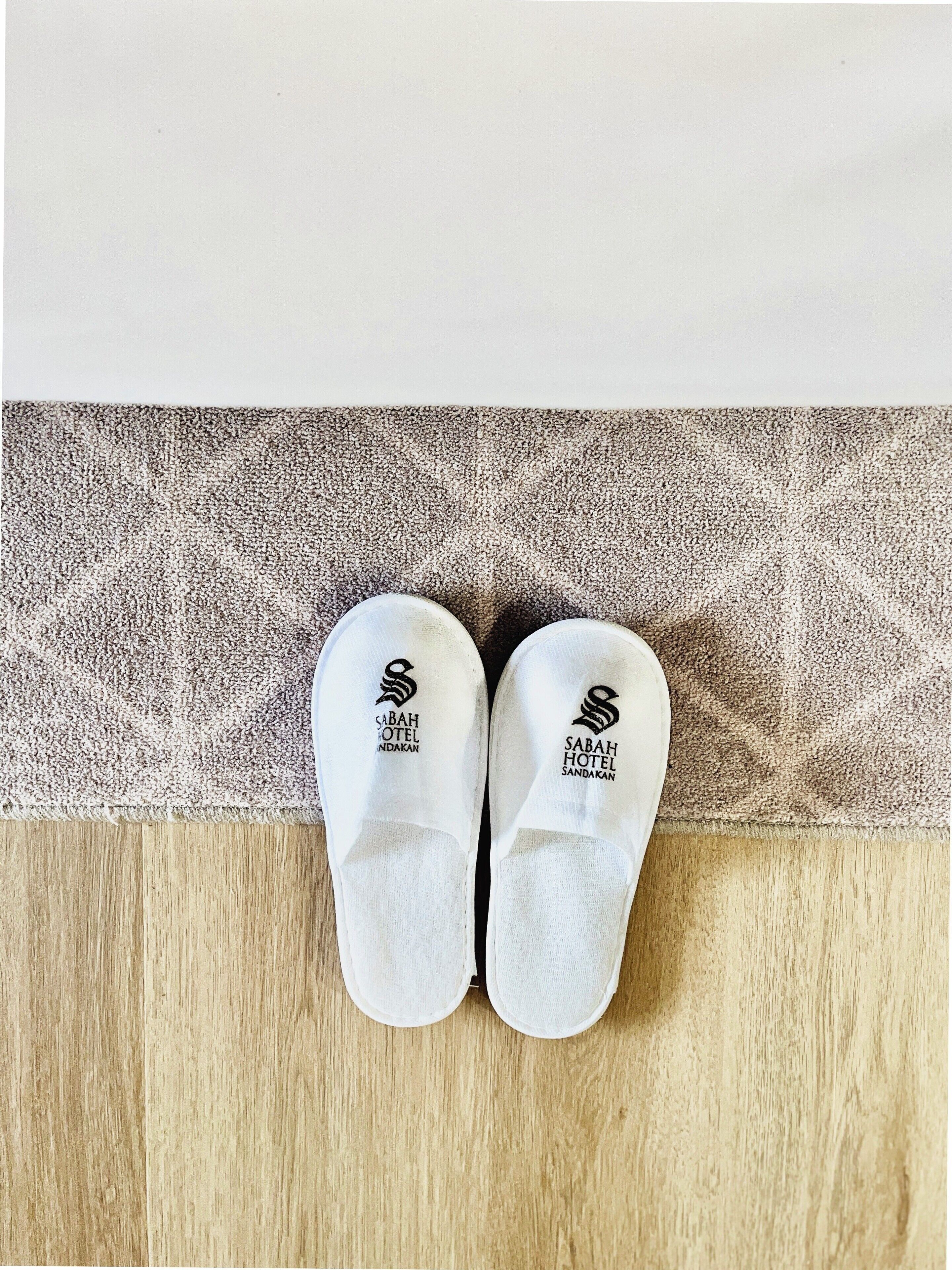 Un par de zapatillas blancas con un logotipo en la suela, sobre un suelo de madera con un tapete estampado al fondo.