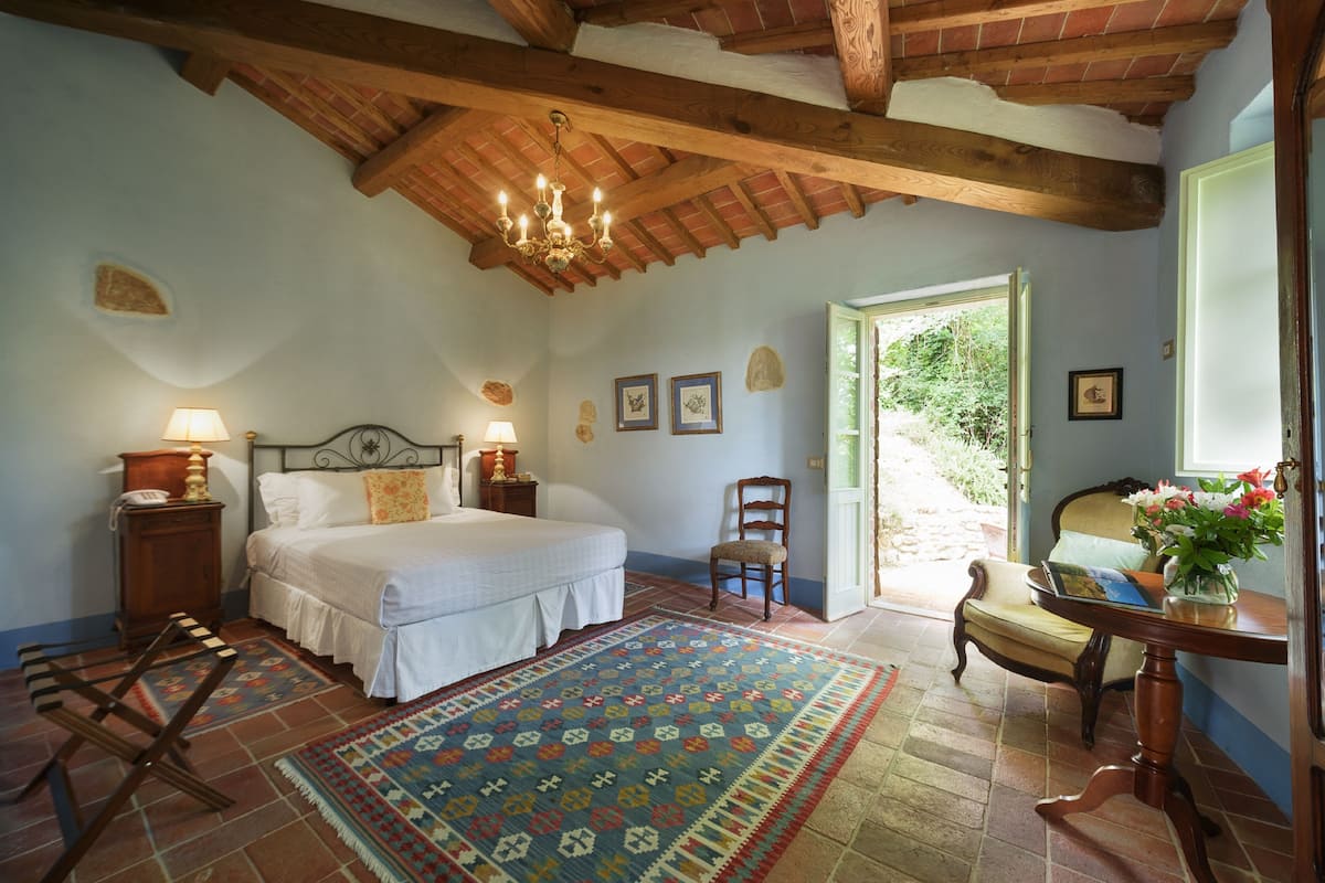 A bedroom with a bed, a chair, a table with a vase, a lamp, and a door leading outside.