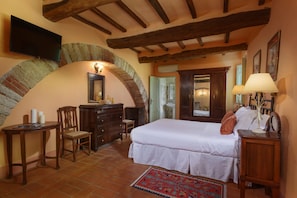 A bedroom with a large bed, a dresser, a chair, a television mounted on a brick wall, and a lamp on a bedside table.