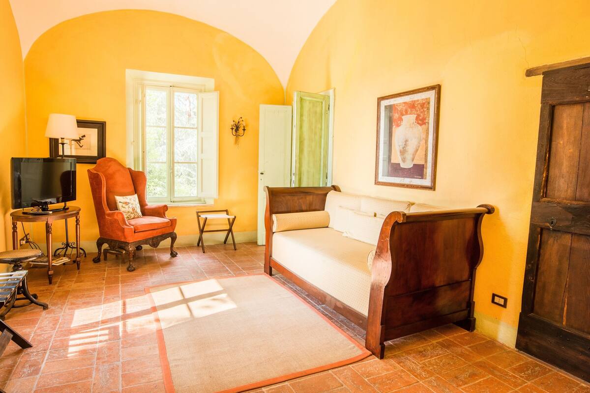 A room with a bed, a chair, a small table, a lamp, and a painting on the wall.