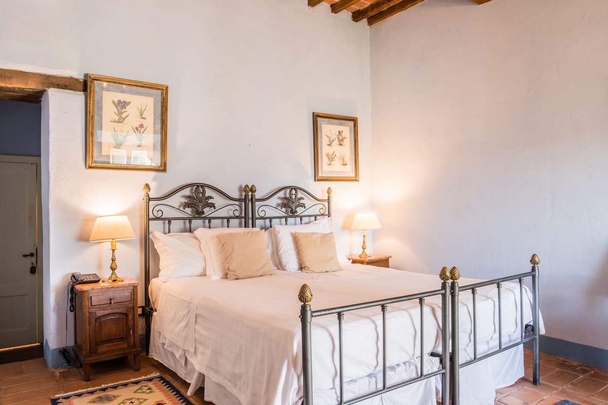 A neatly made bed with a metal frame and white bedding, flanked by two bedside tables with lamps, in a room with white walls and a wooden ceiling.