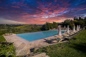 A rectangular swimming pool with a stone deck, surrounded by lounge chairs and umbrellas, set against a backdrop of rolling hills and a colorful sky.