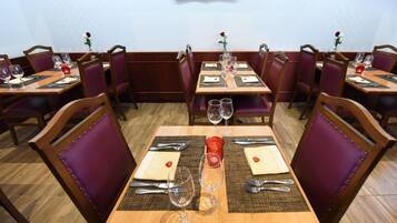 A restaurant with wooden tables and purple upholstered chairs. Each table is set with wine glasses, cutlery, and a red candle.