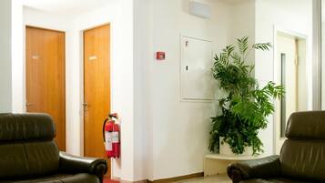 Rebedor amb dues butaques de pell, una taula de cafè de vidre amb revistes, un extintor, una planta en test i portes de fusta amb la indicació "WC" i "RUSH".