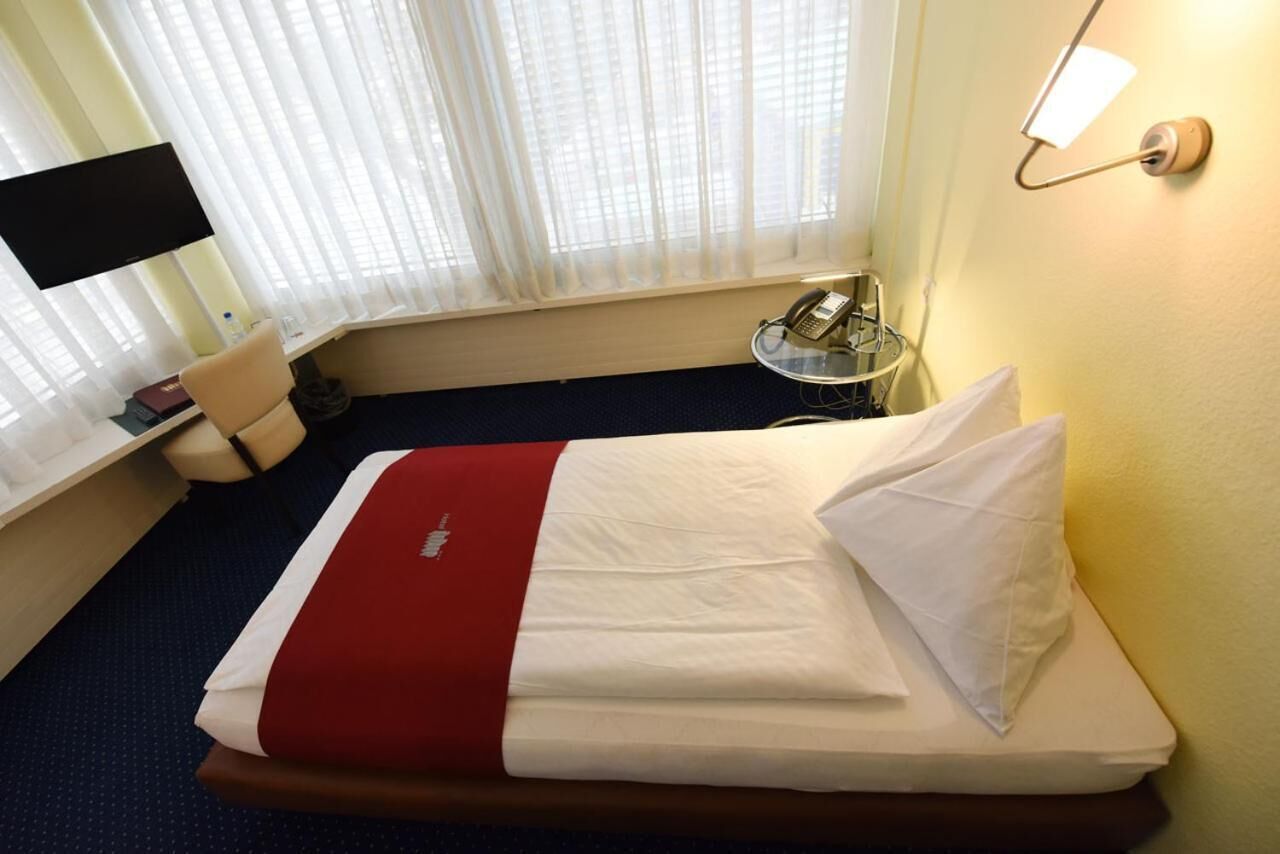 A hotel room with a bed, a desk, a chair, a telephone, and a window with curtains.