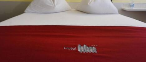 Habitación de hotel con cama, almohadas, colcha roja y mesita de noche con lámpara y botella de agua.