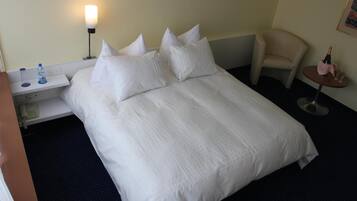 A hotel room with a neatly made bed, a bedside table with a lamp, a chair, and a small table with a bottle and glasses.