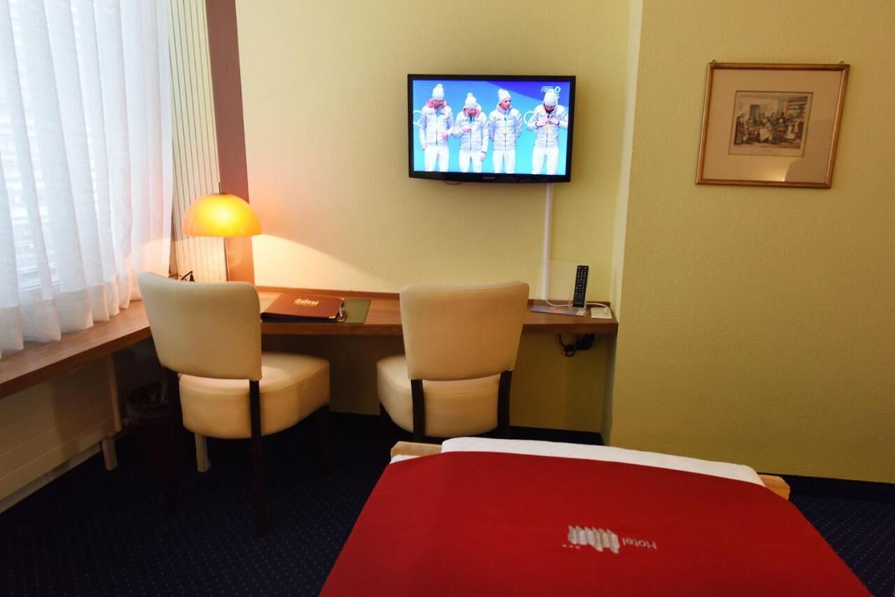Una camera d'albergo con un letto, una scrivania con due sedie, un televisore a parete, una lampada e un quadro incorniciato.