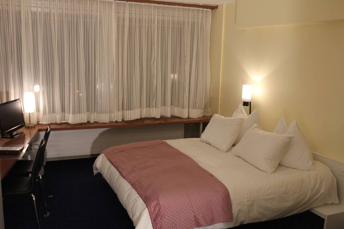 A hotel room with a bed, a desk with a computer, a chair, a window with curtains, and two bedside lamps.