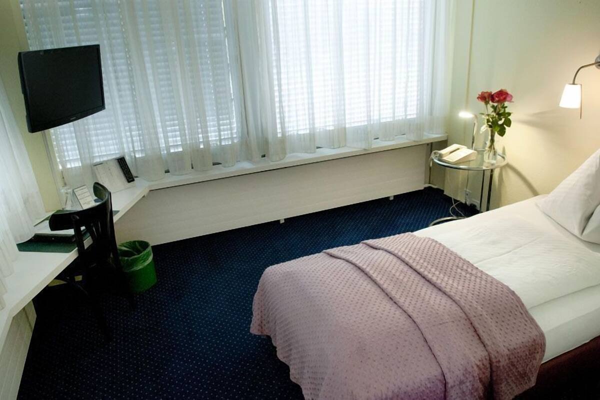 A hotel room with a bed, a desk with a computer, a chair, a TV, a window with curtains, a bedside table with a lamp and a vase of flowers.
