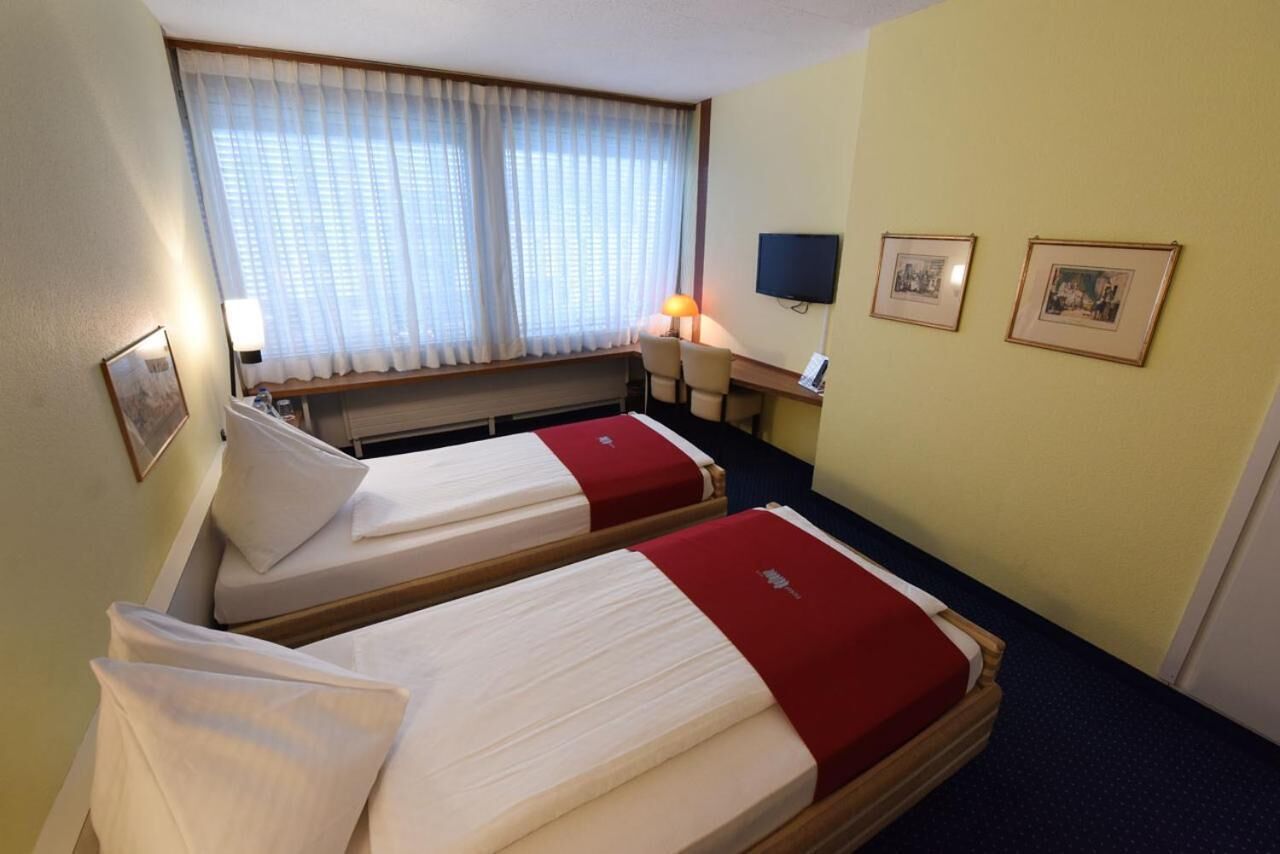 Une chambre d’hôtel avec deux lits simples, un bureau avec une chaise, une télévision et deux tableaux encadrés au mur.