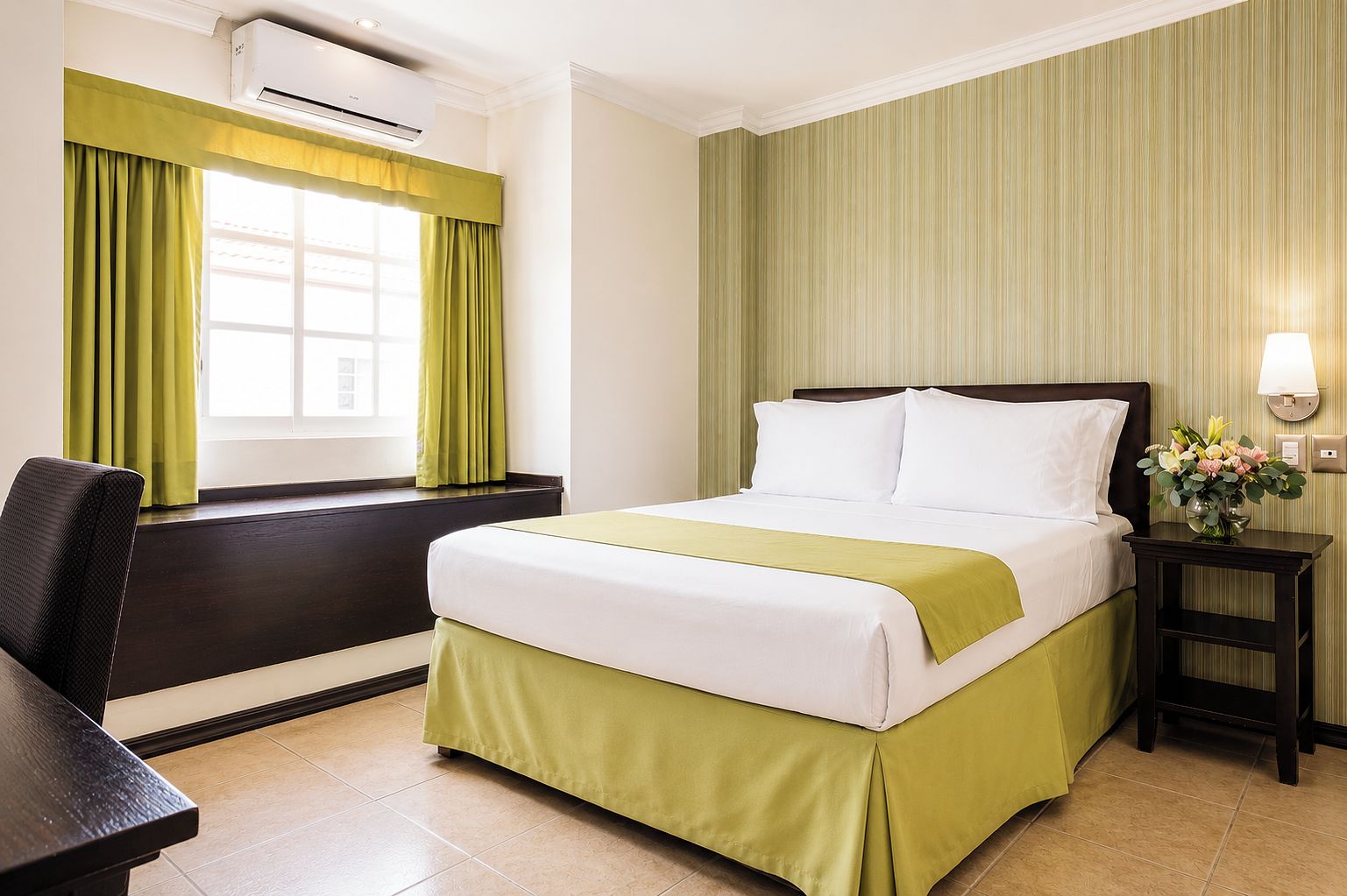 A hotel room with a bed, a desk, a chair, an air conditioning unit, a window with curtains, and a wall-mounted lamp.