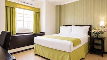 A hotel room with a bed, a desk, a chair, an air conditioning unit, a window with curtains, and a wall-mounted lamp.