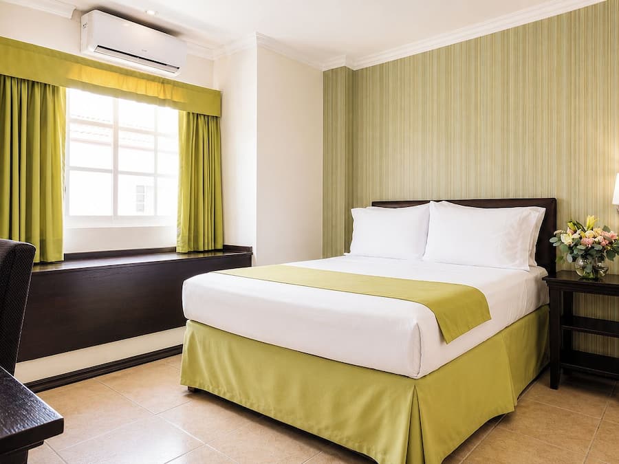 Une chambre d’hôtel comprenant un lit, un bureau, une chaise, un appareil de climatisation, une fenêtre avec des rideaux et une lampe fixée au mur.