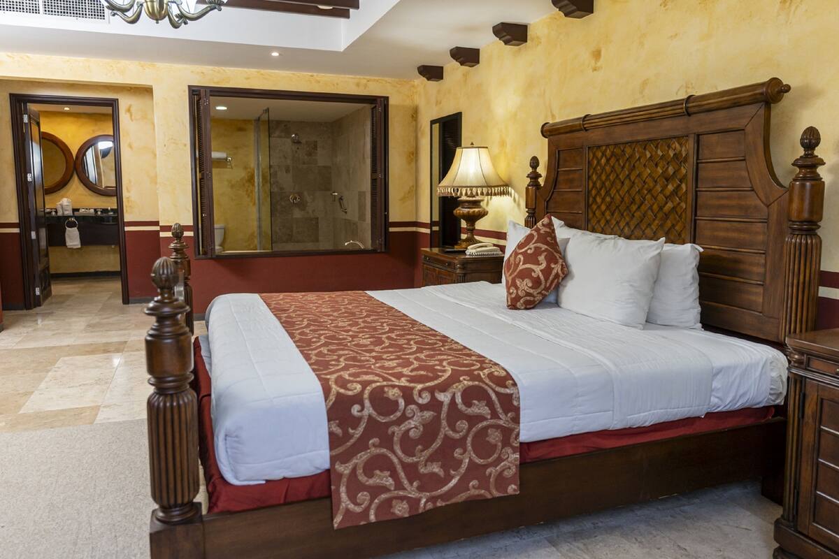 Una camera d'albergo con un letto grande, una testiera in legno, un comodino con una lampada e un bagno visibile tramite una porta aperta.