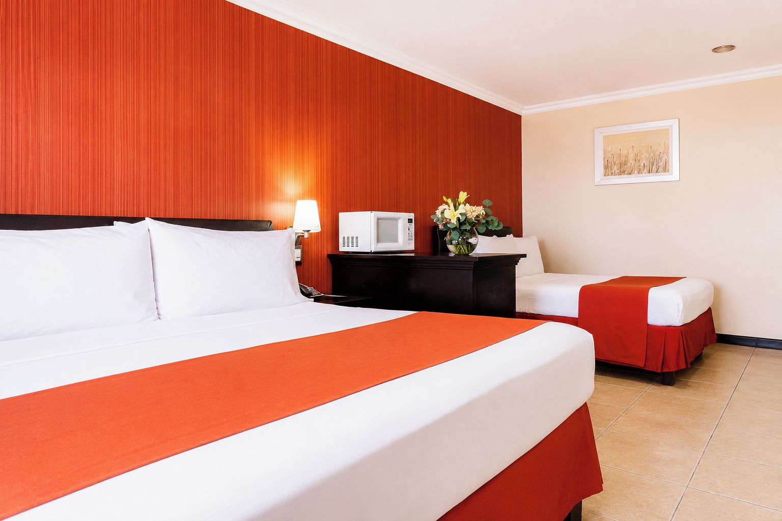 A hotel room with a large bed, a bedside table with a lamp, a microwave, and a small table with flowers.