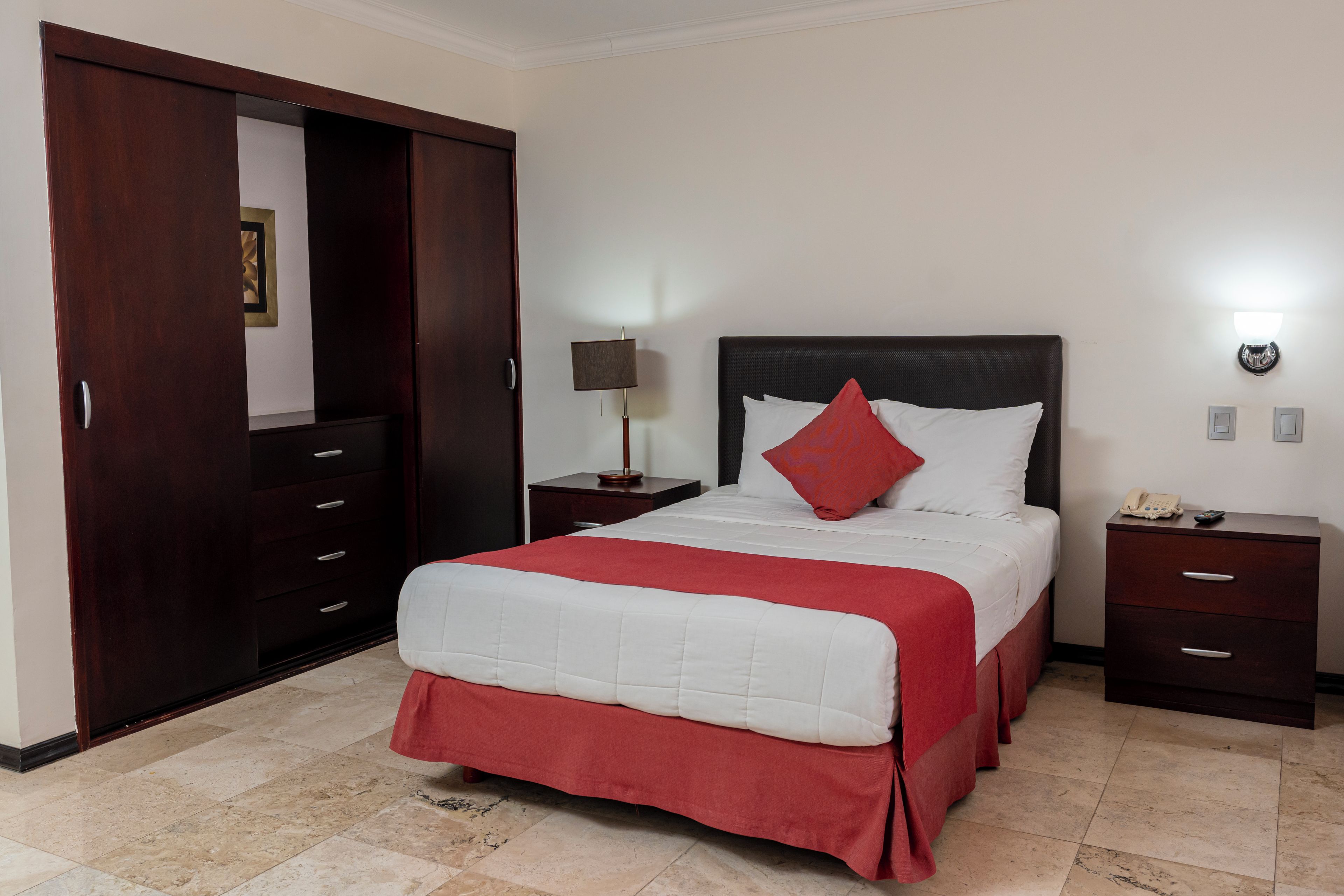 A hotel room with a bed, a nightstand, a lamp, a wardrobe, and a wall-mounted light fixture.