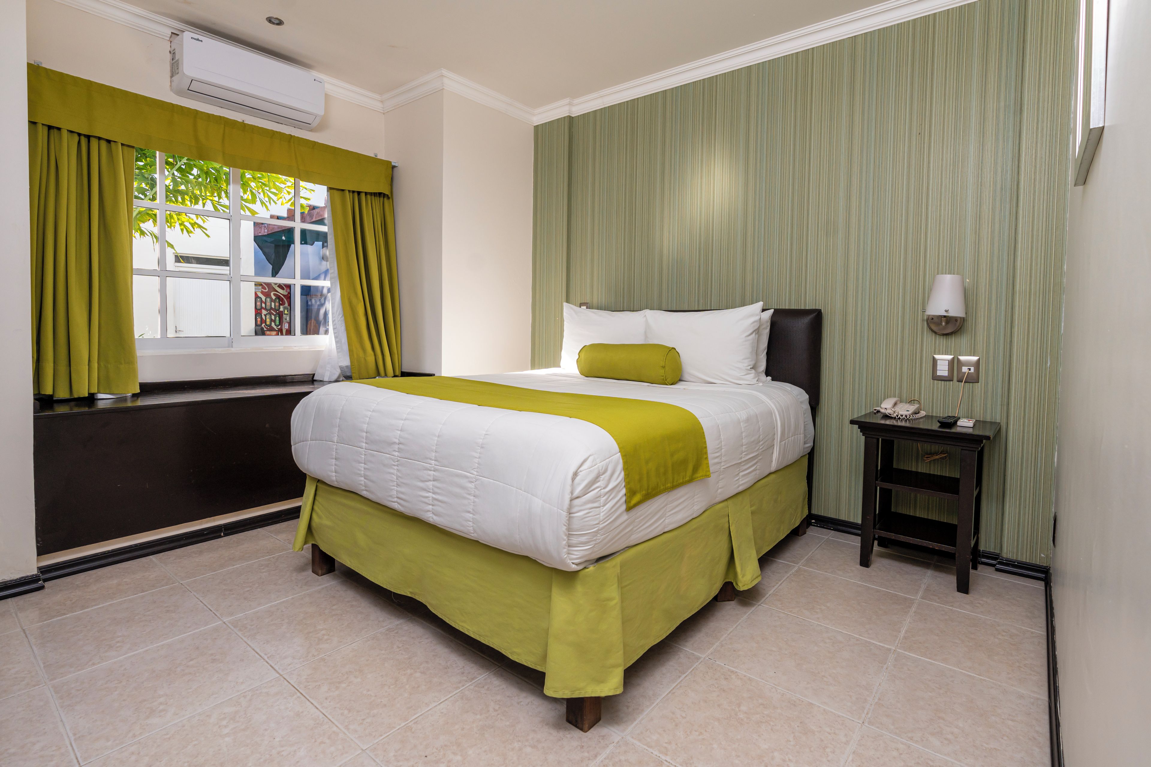 Una habitación de hotel con una cama, una mesita de noche, una lámpara y una ventana con cortinas.