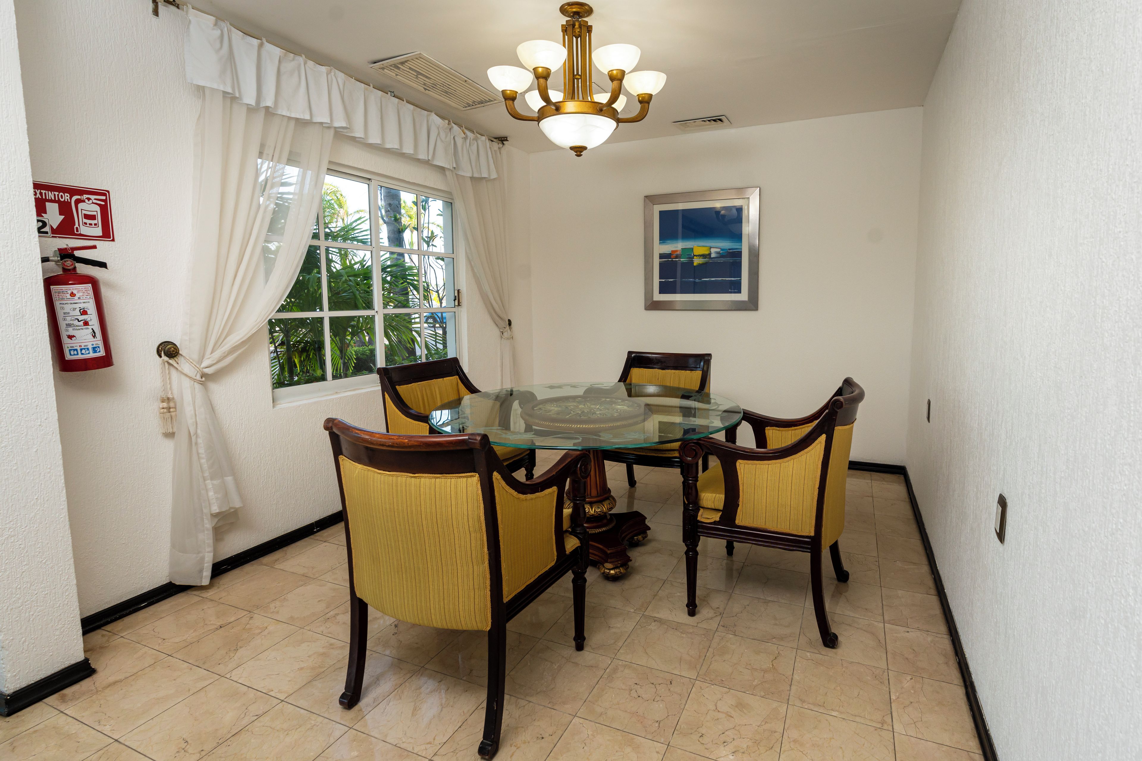 A dining area with a glass-top table surrounded by four chairs with yellow cushions, a chandelier, a fire extinguisher, and a window with curtains.