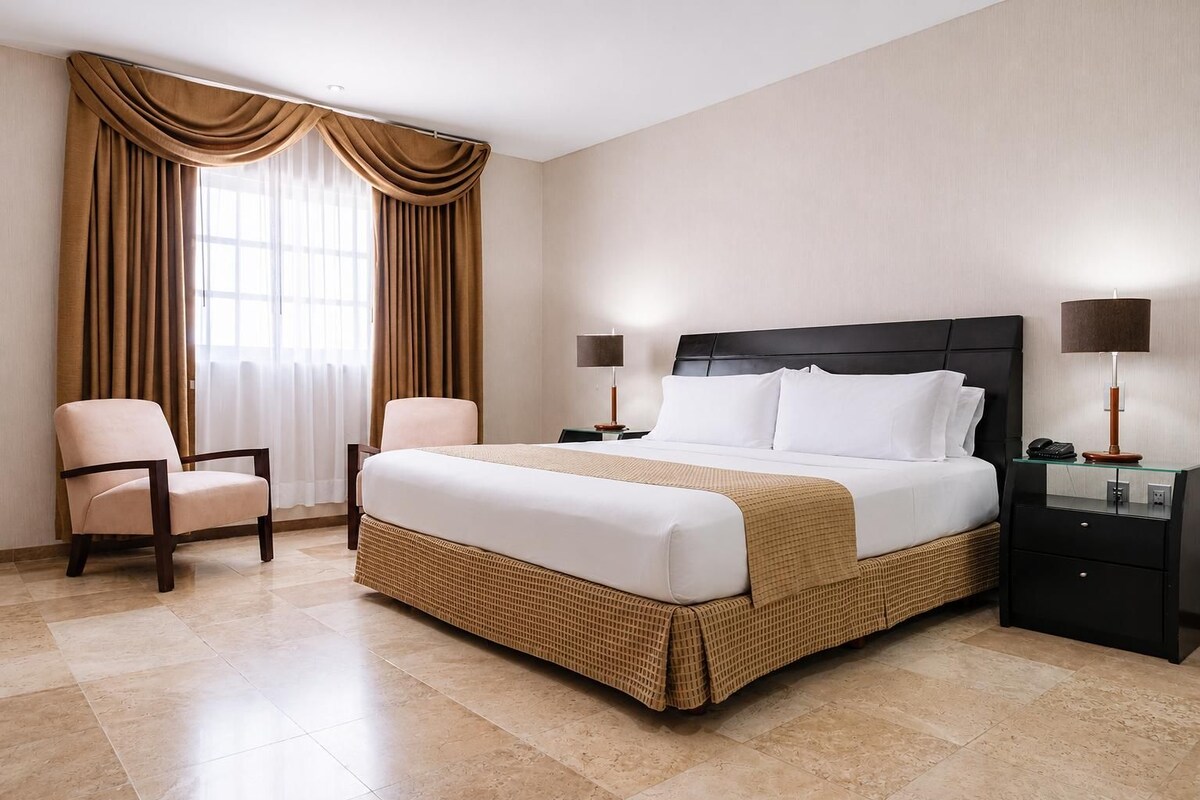 Une chambre d’hôtel avec un grand lit, deux tables de chevet avec des lampes, une chaise et une fenêtre avec des rideaux.