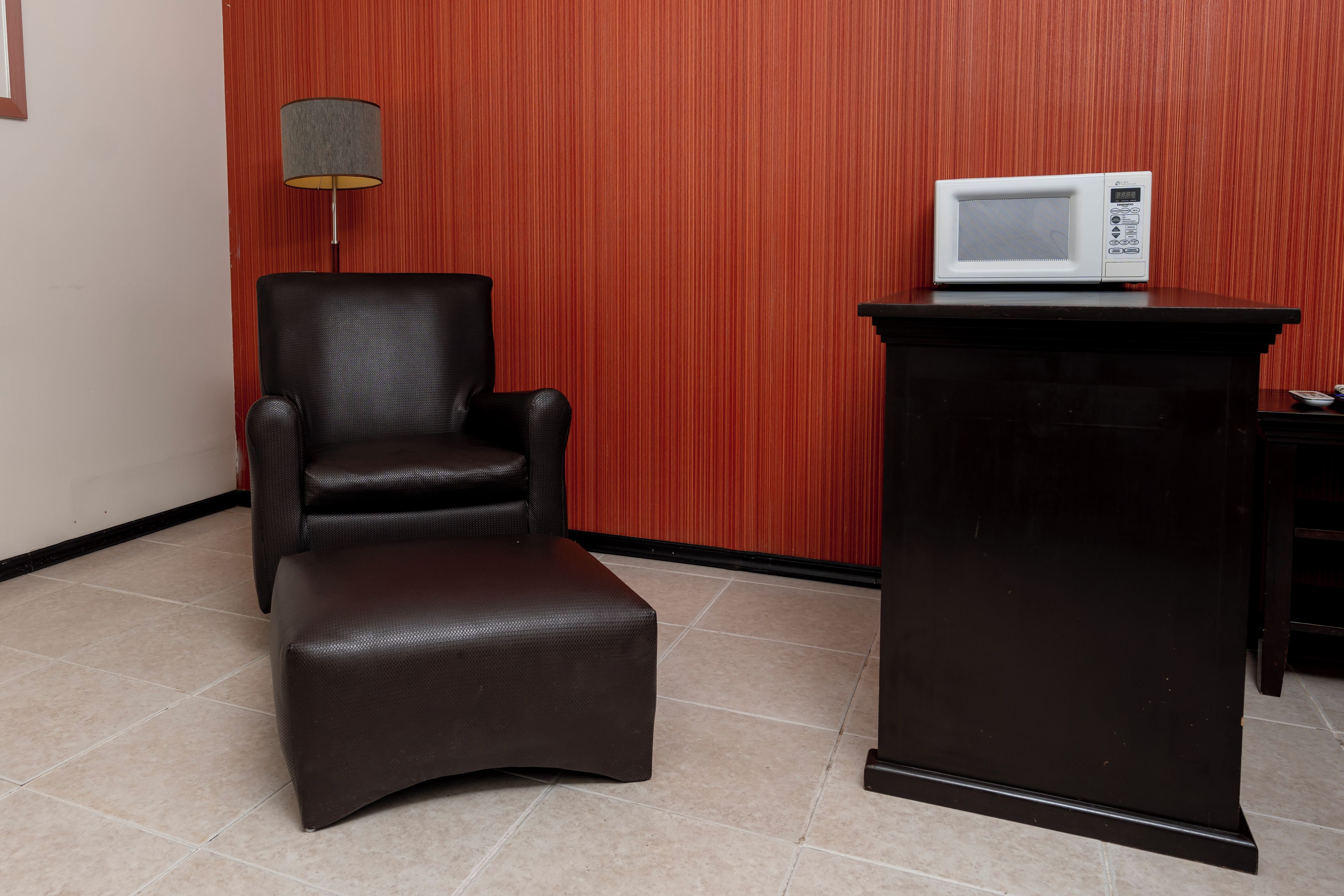 Una habitación con un sillón de cuero marrón, un pequeño otoman, un mueble de madera oscura y un microondas blanco encima.