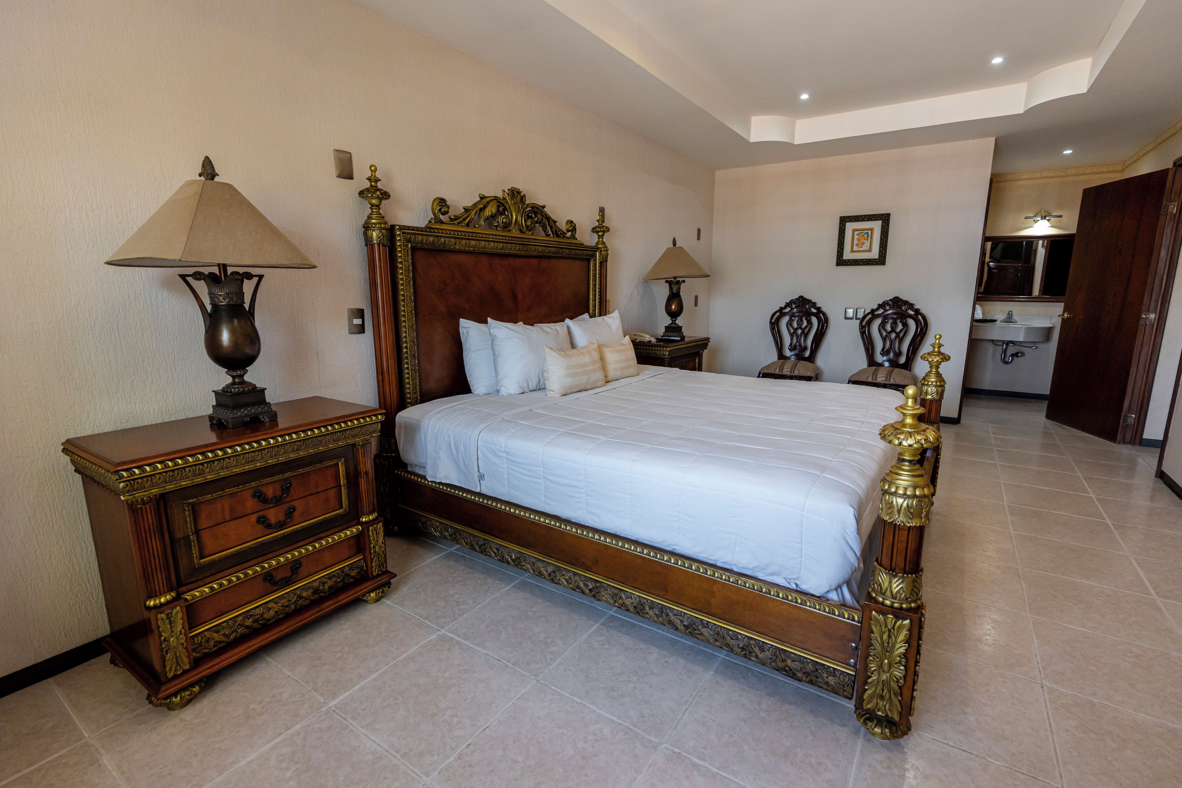 A spacious bedroom with a large bed, two bedside tables with lamps, a wooden dresser, and a bathroom visible in the background.