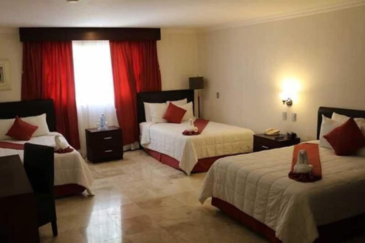 Camera d'albergo con tre letti, tende rosse, un comodino con una lampada e una luce a parete.