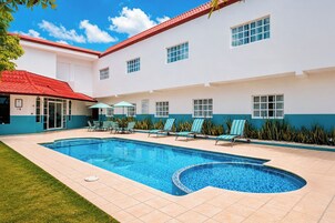 Una piscina con sillas de descanso y sombrillas, rodeada por un edificio de techo rojo y paredes blancas.