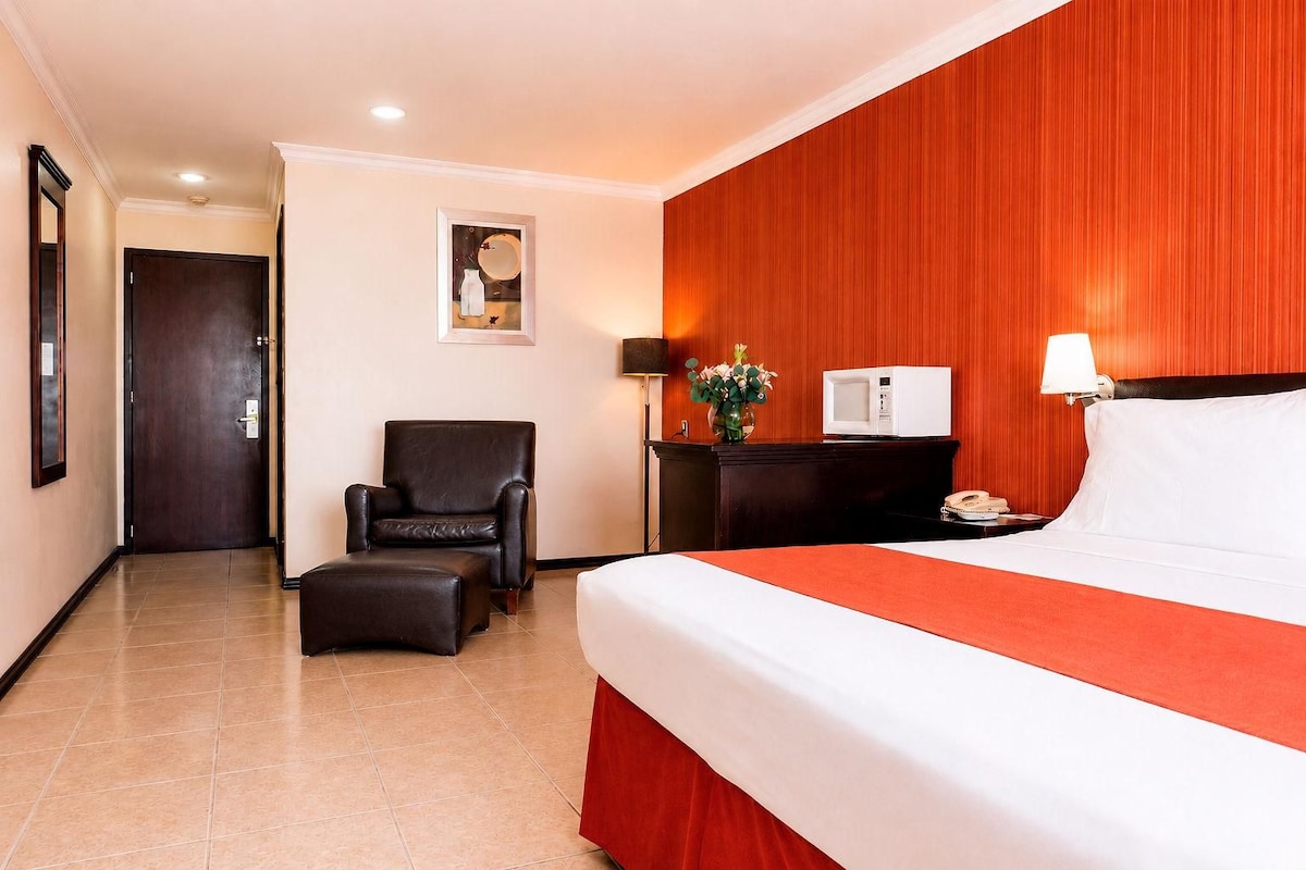 A hotel room with a bed, a chair, a desk with a lamp, and a picture on the wall.