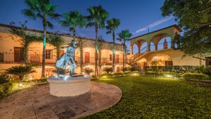 A courtyard with a statue, palm trees, and a building with arches and balconies. - Hotel Posada del Hidalgo by Balderrama Hotel Collection (El Fuerte)