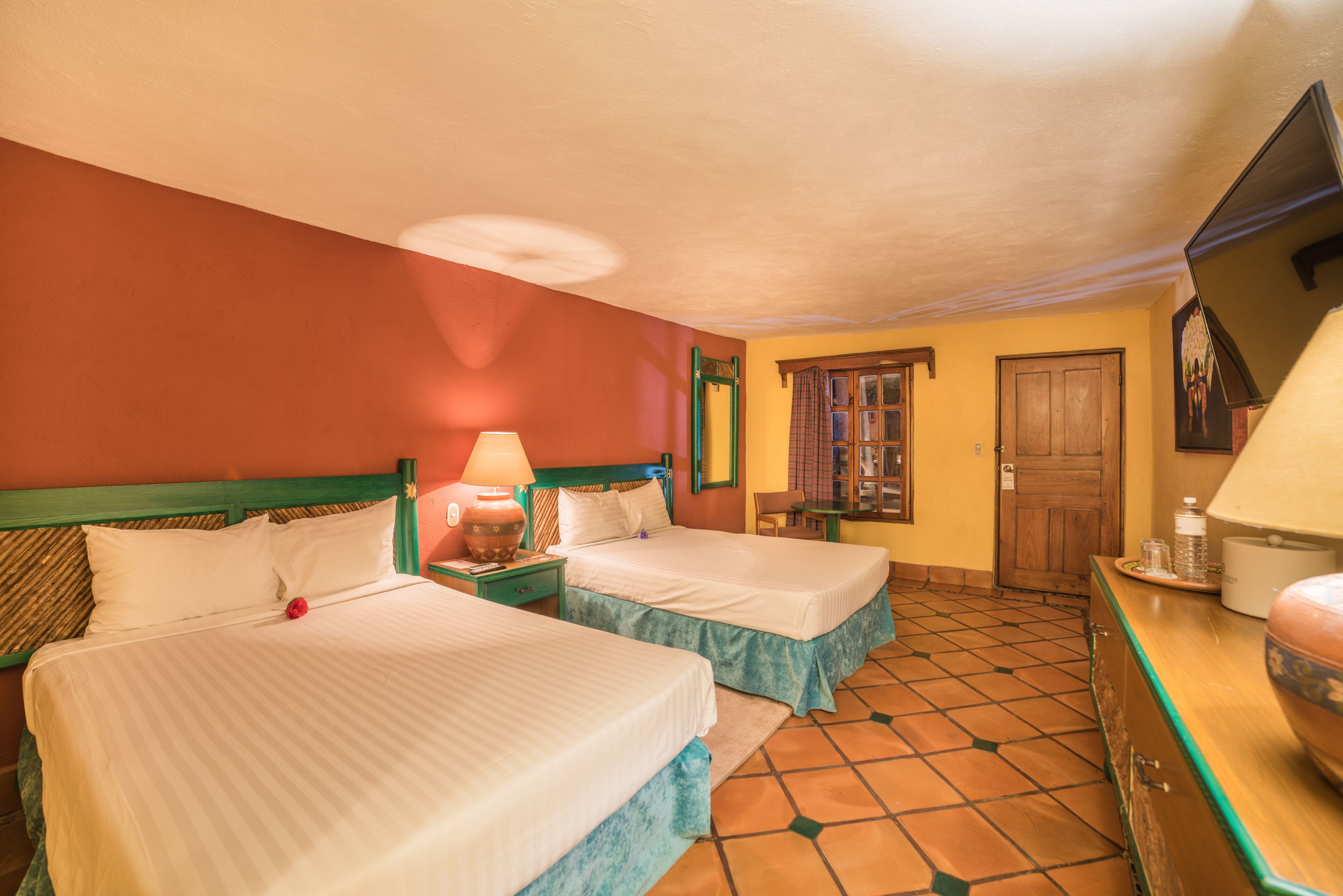 Habitación de hotel con dos camas, una puerta de madera, una mesita con una lámpara y piso de baldosas.