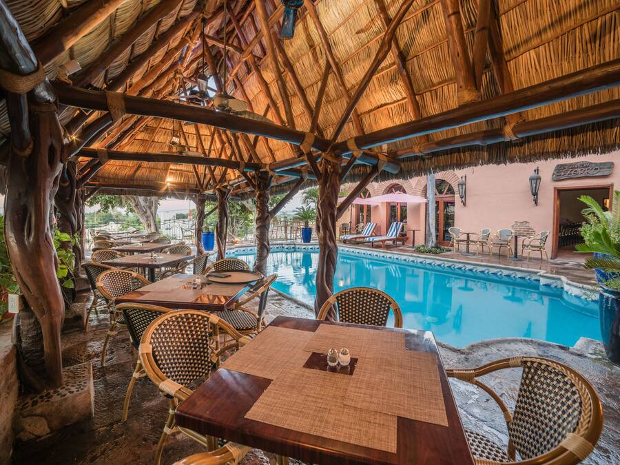 An outdoor pool area with thatched roof seating and tables, surrounded by lush greenery.