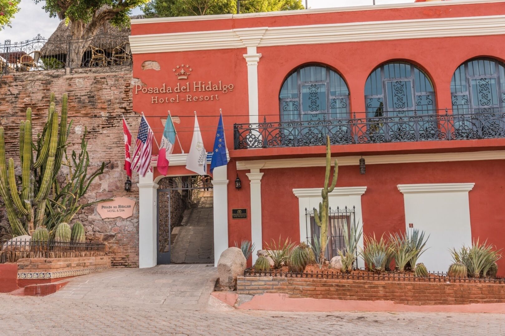 L’entrée de l’hôtel-centre de villégiature Posada del Hidalgo, avec sa façade rouge, ses fenêtres en arc de cercle et son jardin de cactus.