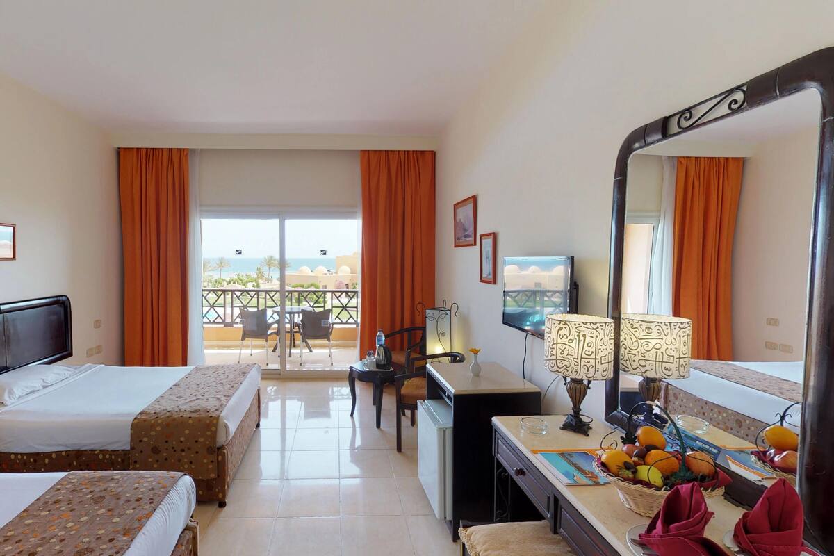 A hotel room with two beds, a desk, a chair, a TV, and a balcony with orange curtains.