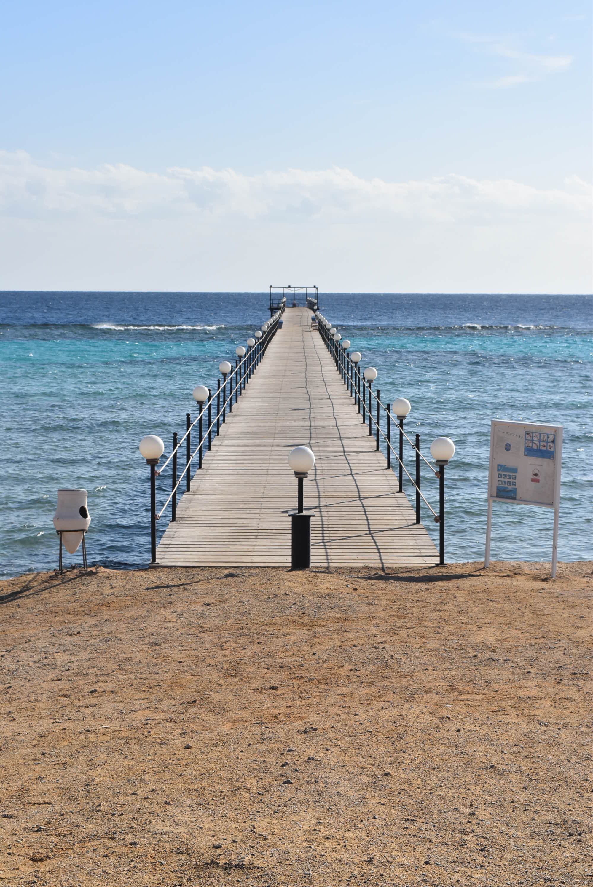 Un muelle de madera se extiende hacia el mar, con barandas y faroles, que conduce a una costa distante.