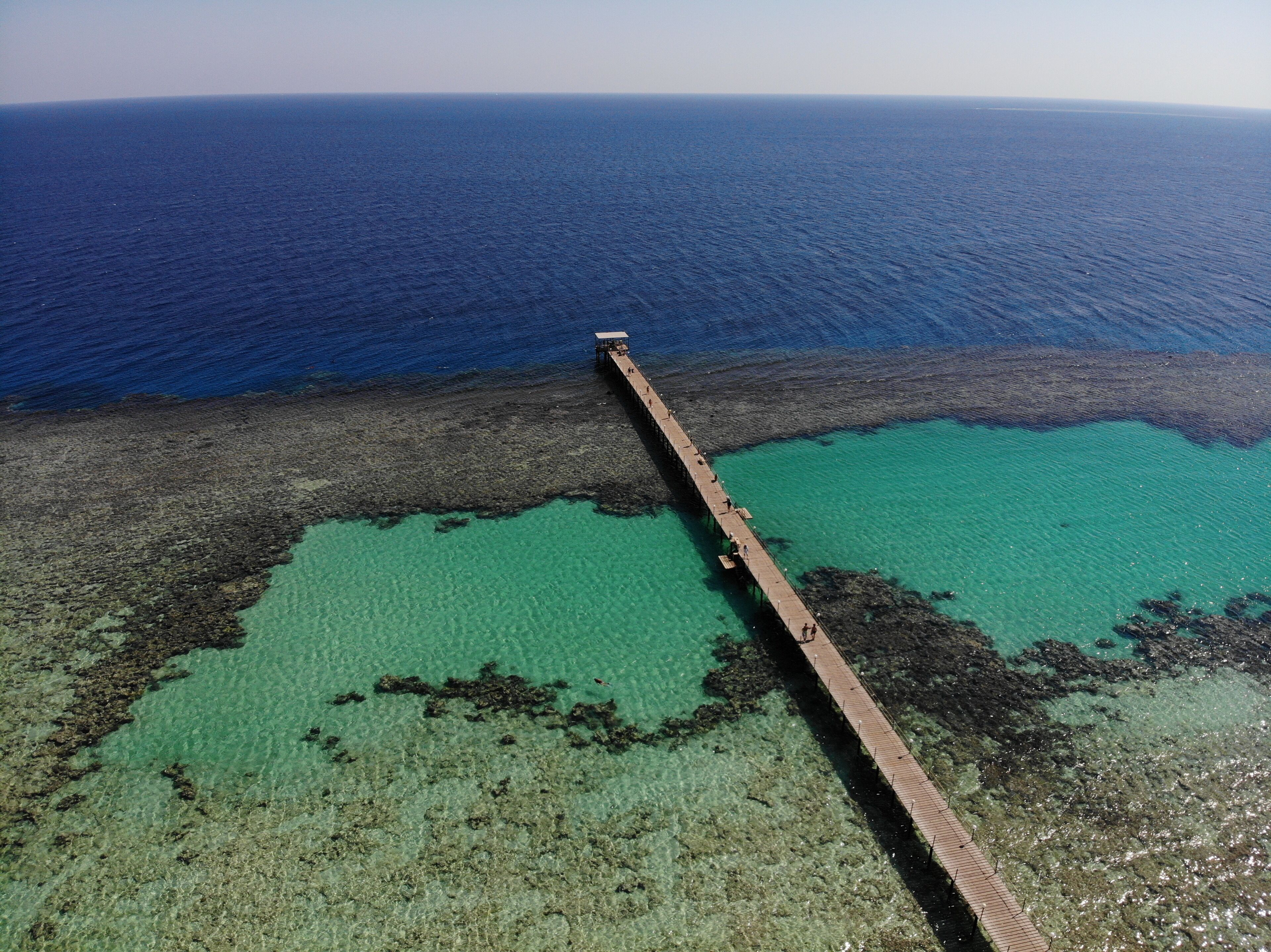 An aerial view of a pier extending into a turquoise sea with clear waters and rocky areas.
