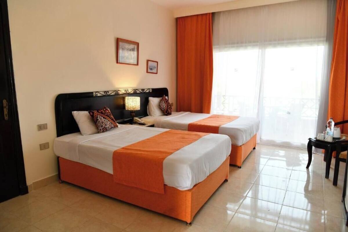 A hotel room with two beds, a nightstand, a lamp, a window with curtains, and a view of the outside.