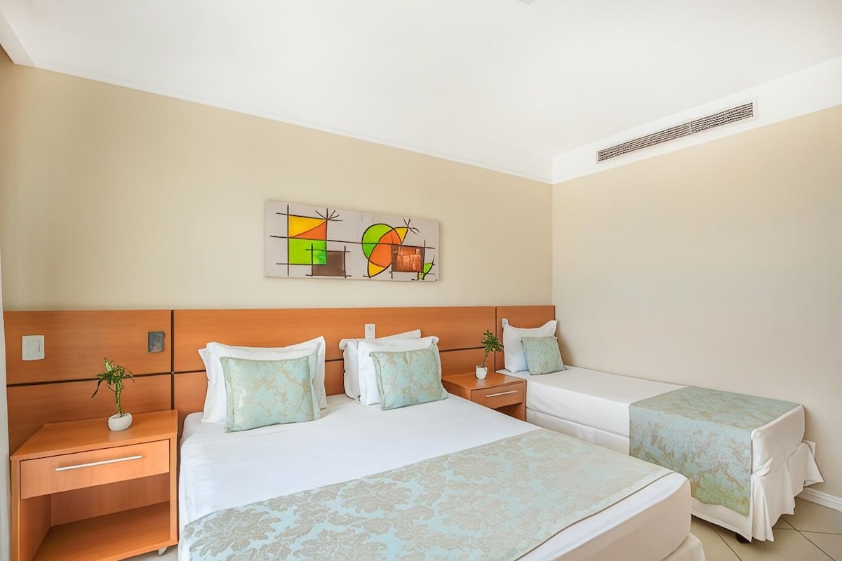 A hotel room with a large bed, two bedside tables, a nightstand, a wall-mounted artwork, and a ceiling vent.