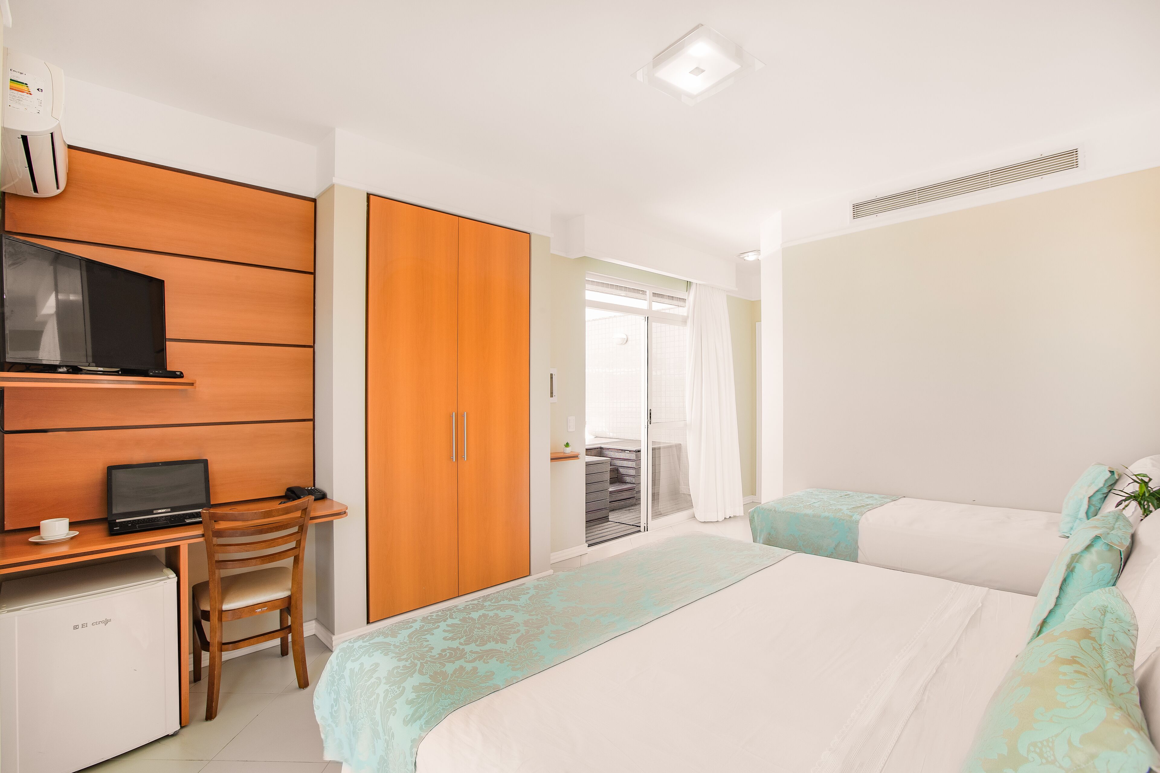 Quarto de hotel com escrivaninha de madeira, laptop, mini-geladeira, cama com roupa de cama estampada e porta de correr para uma varanda.