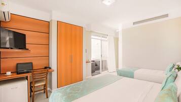 Quarto de hotel com escrivaninha de madeira, laptop, mini-geladeira, cama com roupa de cama estampada e porta de correr para uma varanda.