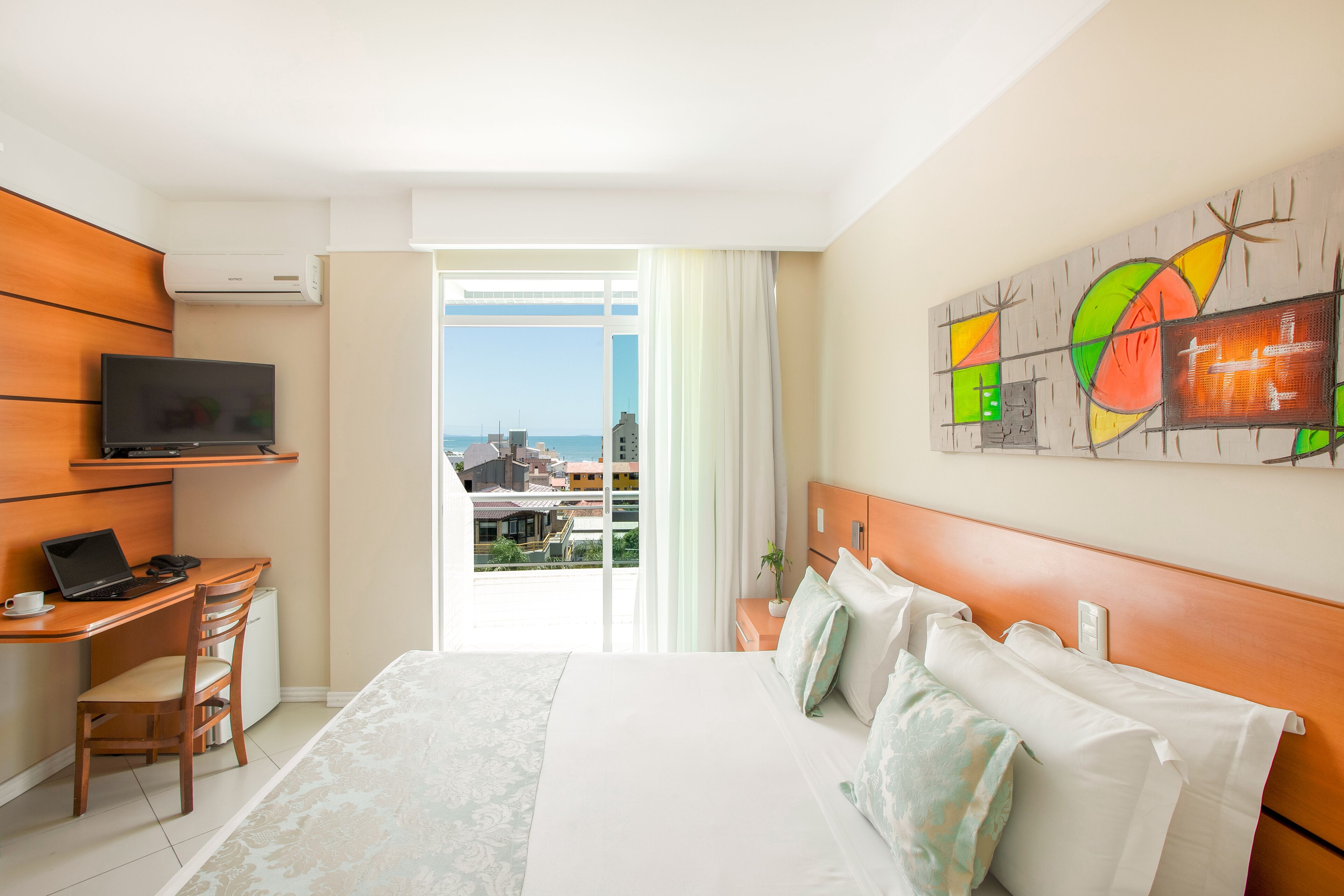 A hotel room with a bed, a desk with a laptop, a television, and a view of the city through the window.