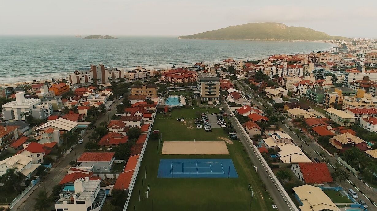 An aerial view of a coastal town with a prominent tennis court, residential buildings, and a beach in the background.