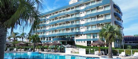 Un hotel de varios pisos con piscina, palmeras y sombrillas de paja.