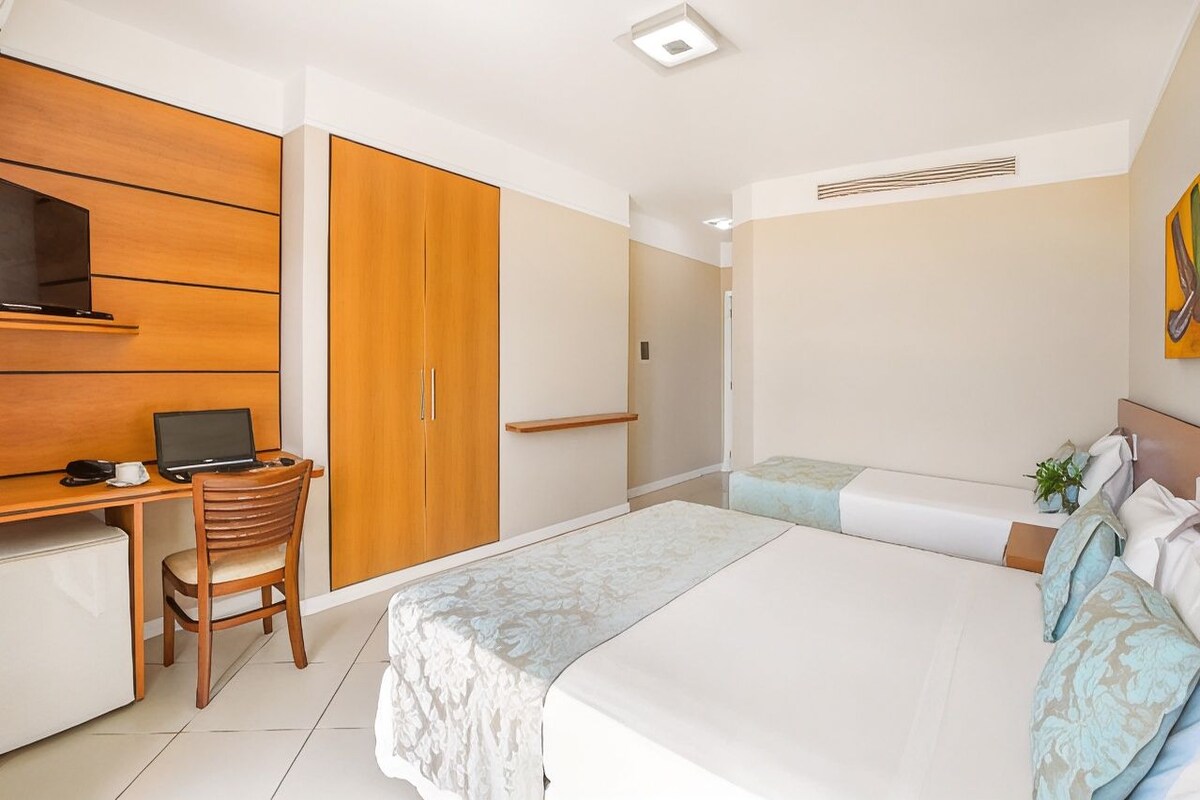 A hotel room with a bed, a desk with a laptop, a chair, a wardrobe, and a small plant.
