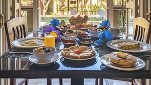 Uma mesa de jantar posta para o café da manhã, com panquecas, frutas e uma tigela de cereal, em um quarto com janelas amplas e vista para um pátio.