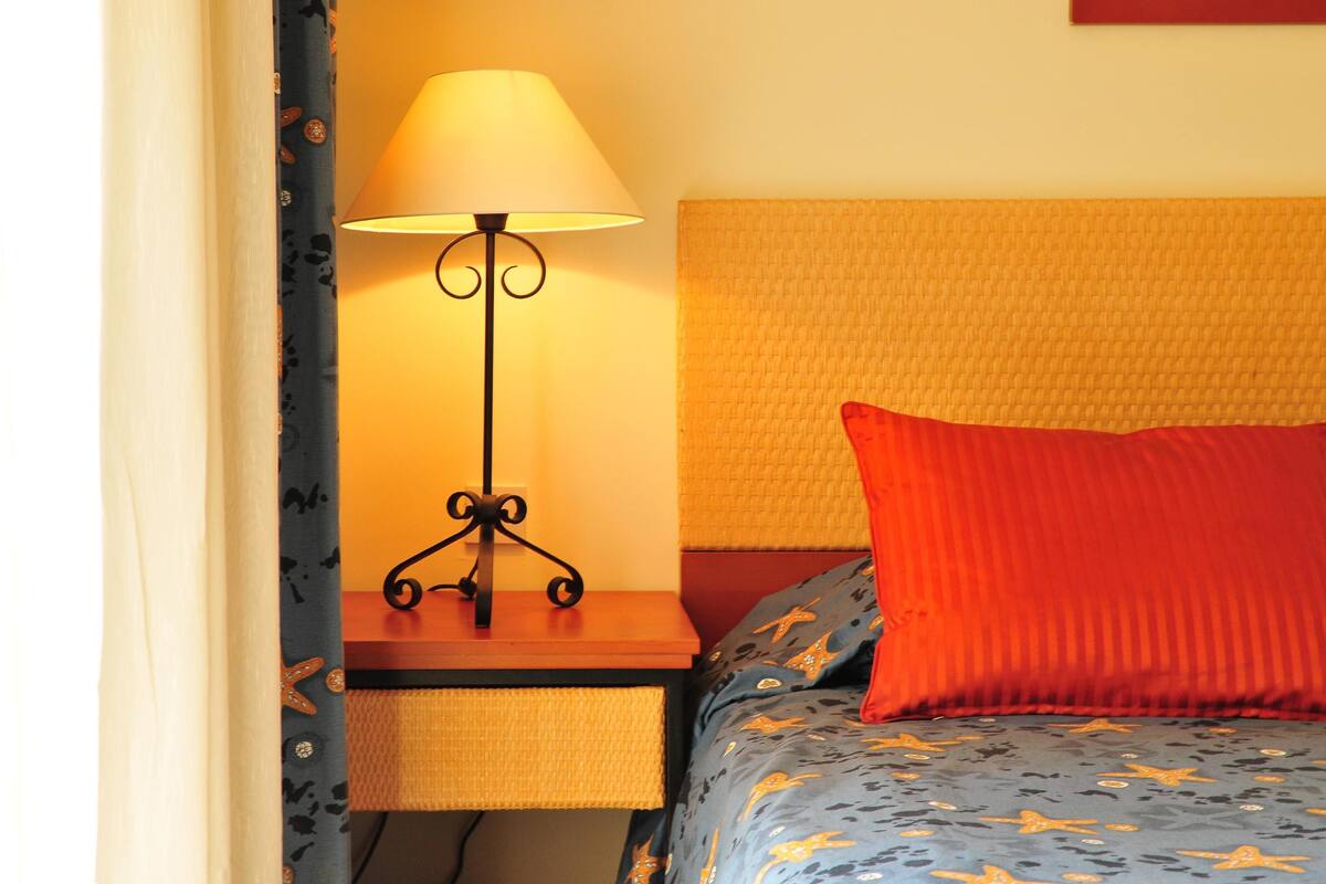 A neatly made bed with a blue and orange patterned bedspread, a red pillow, and a black floor lamp with a white lampshade on a wooden bedside table.